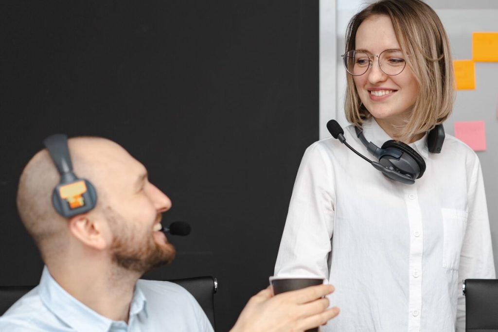 Senior call center agent giving guidance to a junior customer support representative while both smile in office.